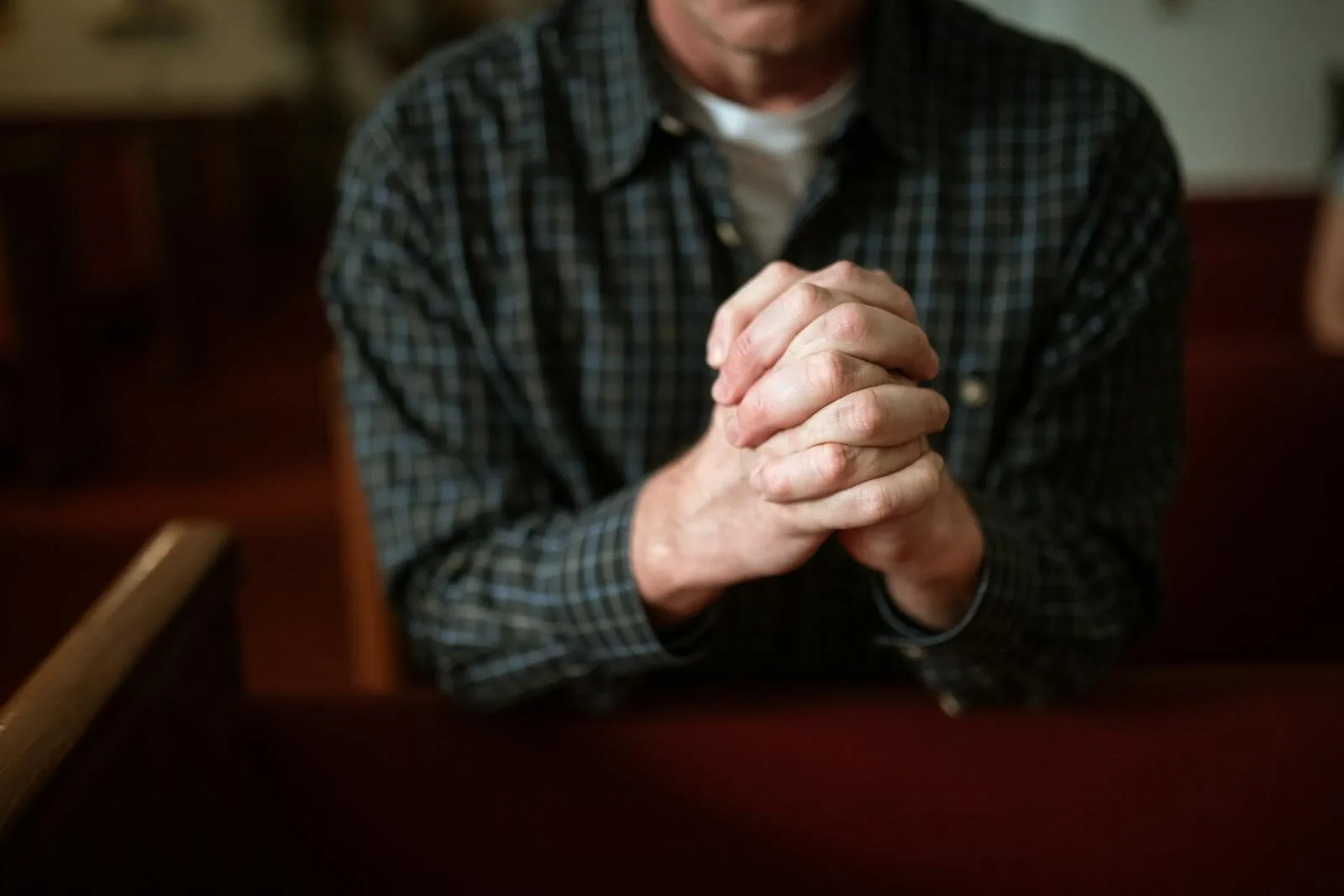 A man in a checked shirt praying in a church, hands clasped together.