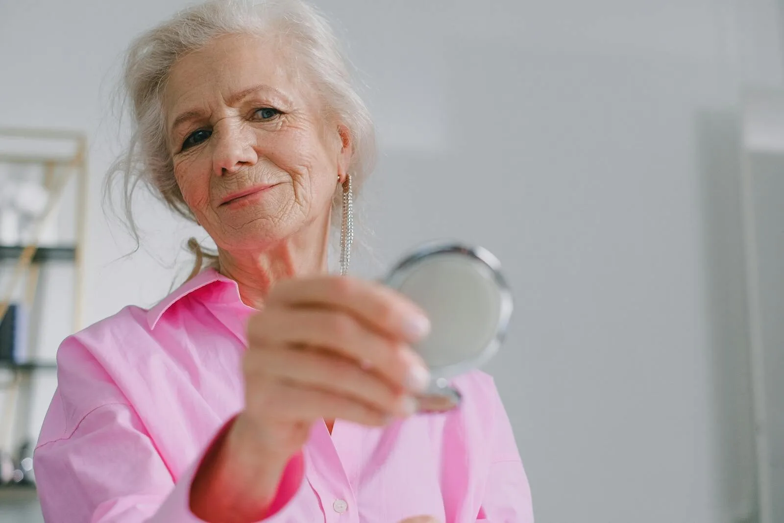 Elegant elderly woman in pink shirt holding a compact mirror indoors, reflecting joy and confidence.