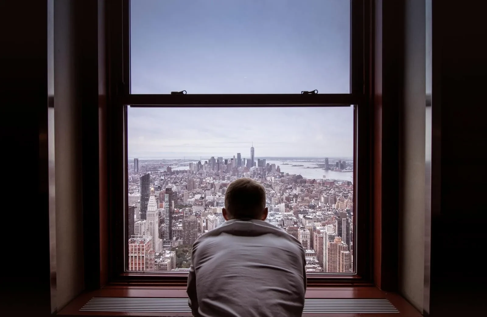 A man reflects while overlooking the iconic New York City skyline through a large window.