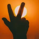 A silhouetted hand forming a peace sign against a vibrant orange sunset in Aaqrabâte, Syria.